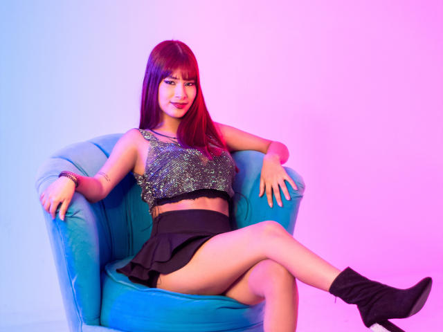 EvilWomen with long red hair, sparkly crop top, black skirt, and black boots, sitting on a blue chair, legs crossed, smiling, pink and blue lighting, sexy.