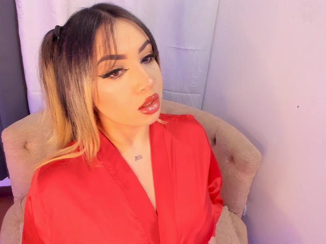 MelissaLust with puffy red lips, dark eyeliner, and blonde hair in pigtails, wearing a red satin robe, sitting in a beige chair, looking at the camera.