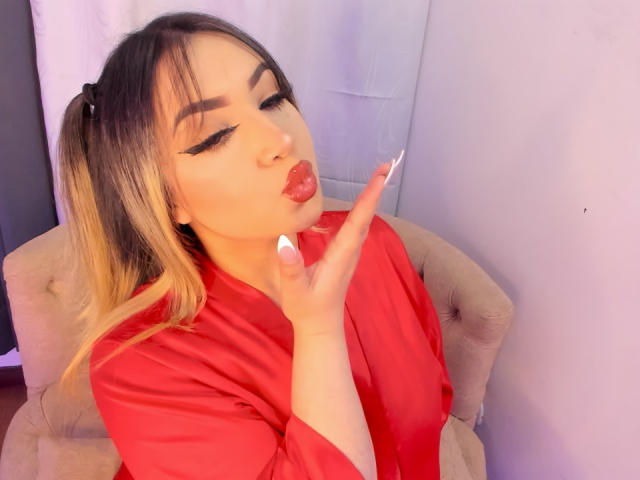MelissaLust con labios fruncidos, camisa roja y uñas blancas, besando sus dedos, cabello rubio en coletas, delineador de ojos negro intenso, pared rosa suave, silla beige..