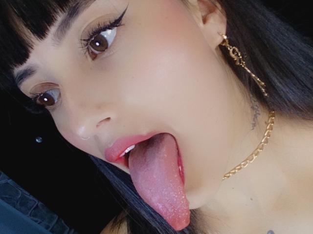 AshlyDeemer's face with tongue out, lips slightly parted, showing pink tongue and small white teeth. She has brown eyes, black eyeliner, and a gold chain necklace.