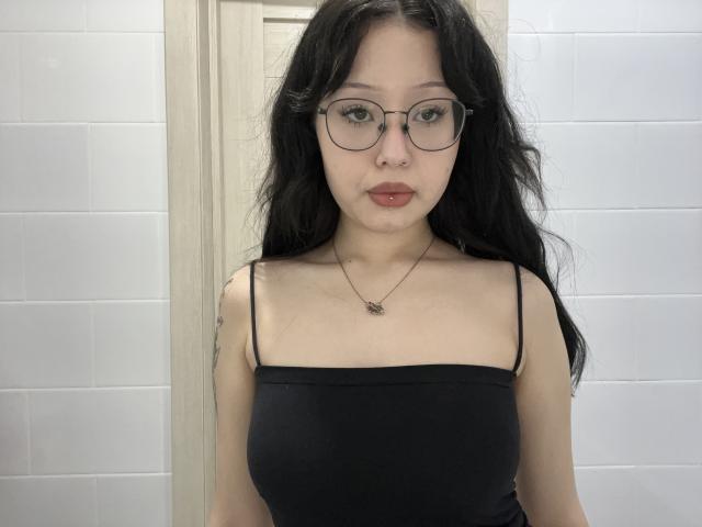 Aretha Assael, con cabello negro largo, gafas y lápiz labial rojo, vestida con una blusa negra de tirantes finos, mostrando pequeños senos, de pie frente a una pared blanca con azulejos..