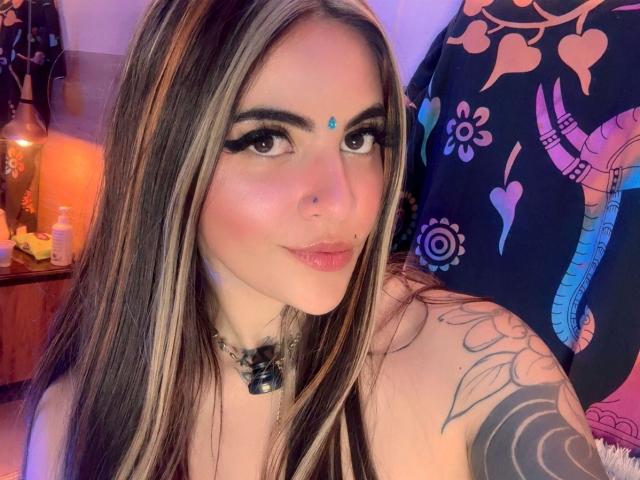 CoriPaige met lang, bruin haar met blonde highlights, een blauwe steen op het voorhoofd, zwarte eyeliner, een neusring, roze lippenstift, een tatoeage op de schouder, een zwarte ketting, lachend, in een slaapkamer..