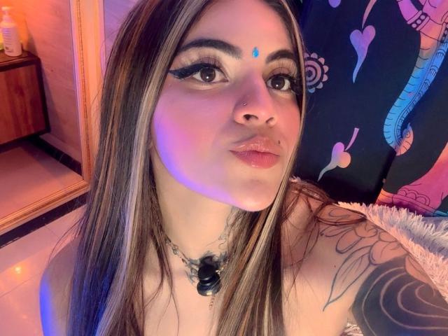 CoriPaige has long brown hair, dark eyeliner, a blue dot on her forehead, a nose ring, and a black necklace. She's pouting with glossy lips and a tattoo on her shoulder.