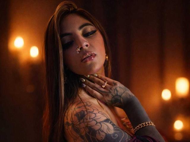 CoriPaige has long brown hair, a nose ring, and tattoos on her arm. She's wearing makeup with dark eyeliner, gold rings, and a bracelet, looking sexy with her hand on her neck.