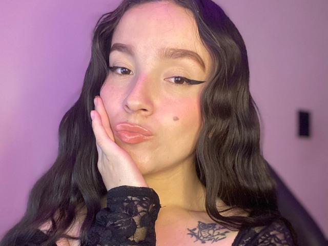 MeliLopezX with pouty lips, black eyeliner, wavy brown hair, lace sleeve, hand on cheek, small mole on cheek, tattoo on chest, pinkish skin, looking at camera.