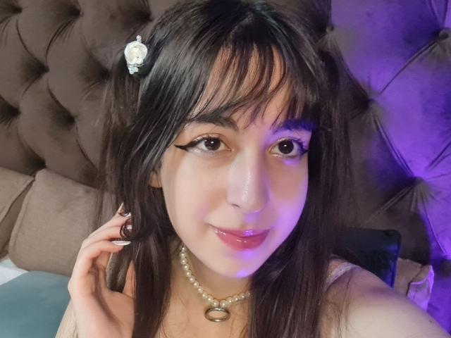 LeiaCrane with dark brown hair, bangs, and a white flower clip, wearing a pearl necklace, has a cute smile, glossy pink lips, and is touching her hair.