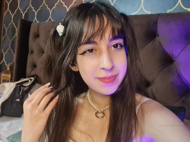 LeiaCrane with dark hair in pigtails, wearing a pearl necklace, has eyeliner, pink lipstick, and is touching her hair, sitting on a bed with a tufted brown headboard.