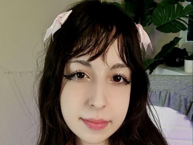 LeiaCrane has pale skin, dark brown wavy hair with pink bows, and thick black eyelashes. She's smiling with pink glossy lips. Her eyes are dark and she's looking at camera.