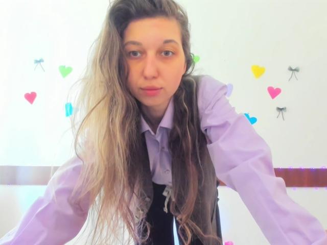Sanella with long, wavy brown hair, wearing a light purple shirt and gray vest, leaning forward with a slight pout, looking into the camera, showing her cute face.