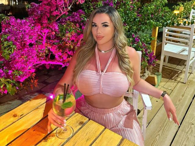 EvaSunderland met grote borsten, lang golvend blond haar, een roze topje met halter, een roze plooirok, die een drankje vasthoudt met groene en roze garnering, zittend aan een houten tafel..