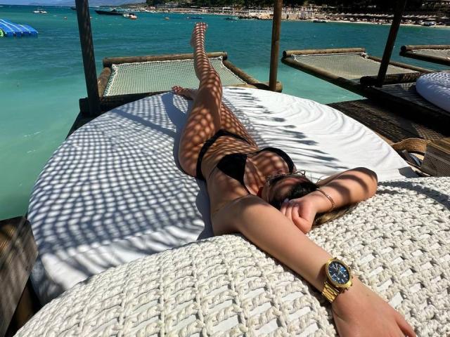 EvaSunderland lying on a bed with a white woven cover, wearing black bikini, legs up, shadowed by wooden roof, blue-green ocean, wristwatch, sunbathing, relaxed, legs spread.