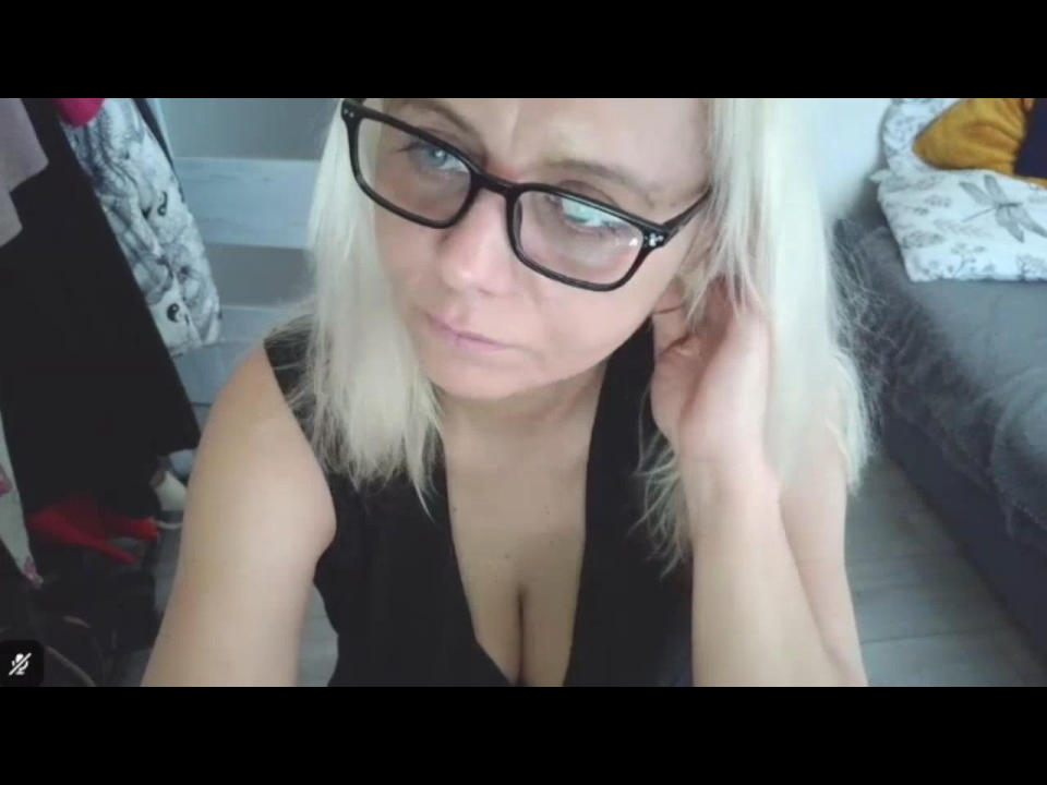 ScarlettWhisper with blonde hair, black glasses, and a black sleeveless top showing her tits. She's looking up with a slight frown, touching her ear with her right hand.