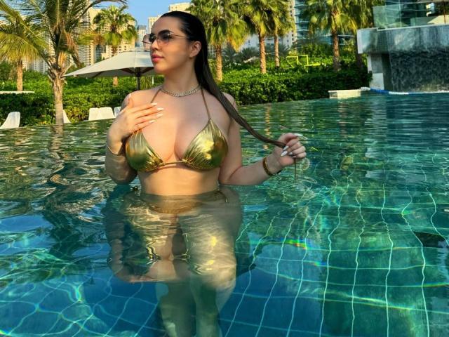EvaSunderland in a green-tinted bikini, standing in a pool, holding her long brown hair, with large tits, wearing sunglasses, and a necklace, with her hand on her chest.