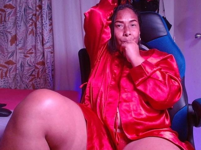 HannaPasion, vestida con una bata roja de satén brillante, está sentada con un brazo levantado, mostrando su zona púbica, y su mano cerca de la boca, en una silla azul, luciendo segura de sí misma..