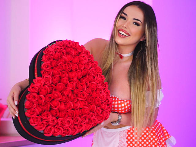 EvaSunderland with long blonde hair, red checkered top, white choker, holding heart-shaped box of red roses, smiling, pink and white background. Boobs visible, cute, sexy.