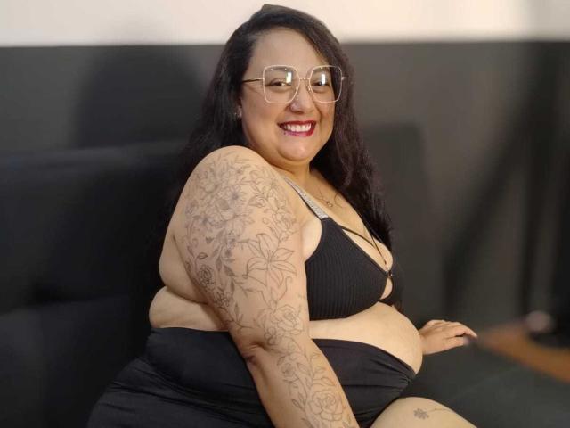 AvaRyd, a plus-sized woman with tattoos on her arm, smiling, wearing glasses, black bra, and black skirt, sitting on a dark sofa, showing her belly and tits.
