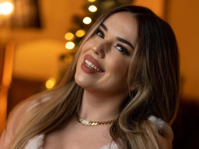 EvaSunderland with long blonde hair, smiling at the camera, wearing a white top and gold necklace, her tits partially visible, warm lighting, bokeh background, soft smile.