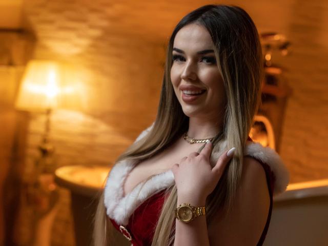 EvaSunderland with long brown hair, smiling, wearing a red velvet dress with white fur trim, showcasing her big boobs, gold watch, and necklace, in a warmly lit room.