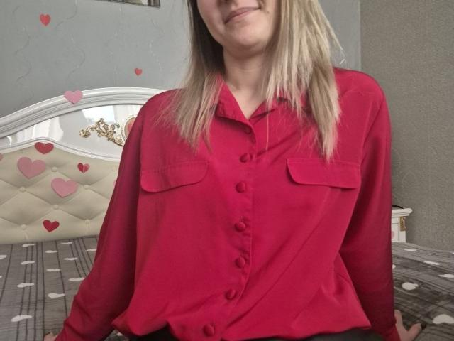Anshante with blonde hair, wearing a red button-up shirt with chest pockets, sitting on a bed with heart decorations, smiling slightly. Boobs not visible, ass not visible.