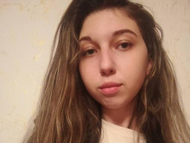 Close-up of Sanella with long, slightly messy brown hair, light brown eyes, and neutral expression. She has fair skin, small nose, and slightly parted pink lips. Sanella wears a simple white top. Her hair has subtle blonde highlights. She has small gold earrings. The image has a soft, warm light, highlighting her facial features. The background is a plain, slightly textured wall. The overall composition is centered and slightly cropped.