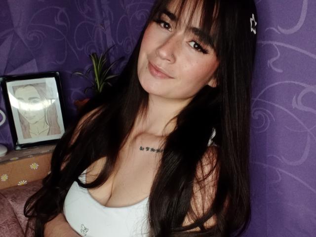 EllieBonneth with long black hair, white top showing her tits, subtle smile, nose ring, tattoo above chest, purple wall, framed picture, plant, and pink cushion in background.