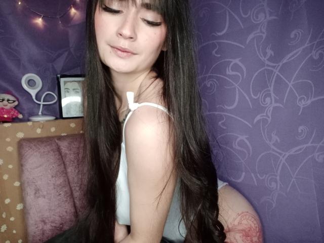 EllieBonneth with long brown hair, eyes closed, smiling slightly, wearing a white tank top, showing her white-pussy and tattoo on her right ass-cheek, in a purple room.