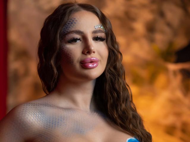 AshleySinnX with wavy brown hair, blue scale paint on face and shoulders, glossy pink lips, and a diamond hair clip, showing off her tits in a sexy, detailed shot.