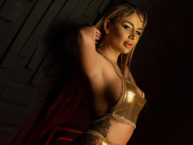 AshleySinnX with gold chain top, exposing her tits, has a tattoo on her side, and a red velvet cape, posing seductively with one arm raised, dark background.