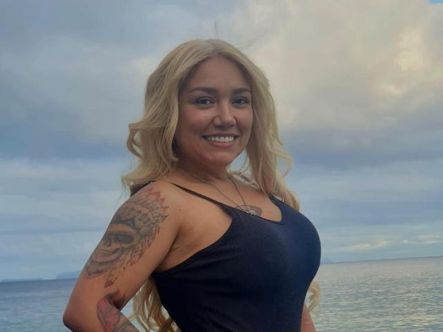 Sonia Ferrer, mit langen blonden Haaren, lächelnd, trägt ein schwarzes Tanktop und zeigt ein Tattoo auf ihrem rechten Arm. Sie steht am Meer, wobei ihre Brüste sichtbar sind, aber die Brustwarzen nicht..
