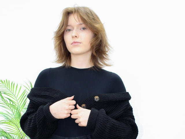 YukiKendal with shoulder-length brown hair, wearing a black knitted sweater, buttoning it, white background, green palm leaves on the left.