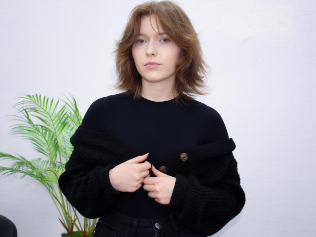 YukiKendal with shoulder-length brown hair, wearing a black sweater, buttoning it, has pale skin, pink lips, and a neutral expression, with a green plant on the left.