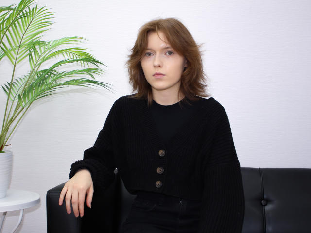 YukiKendal with brown wavy hair, wearing a black rib-knit sweater with buttons, sitting on a black leather couch, arm resting on the seat, green plant to the left.