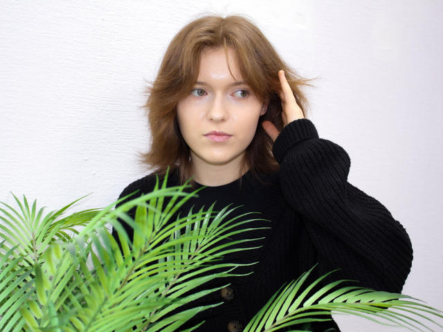 YukiKendal has shoulder-length brown hair, wearing a black knitted sweater, hand in hair, green palm leaves in front, white textured wall behind, serious expression, blue eyes, fair skin.