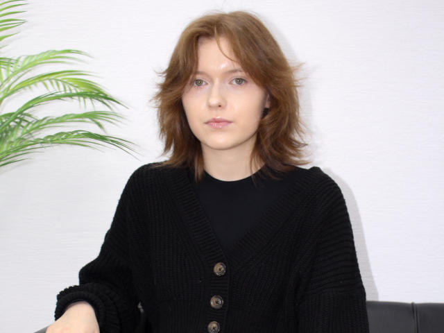 YukiKendal with medium brown hair, wearing a black cardigan with buttons, black shirt, and a neutral expression, next to a green plant. She's got a cute face.