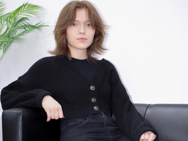 YukiKendal with shoulder-length brown hair, wearing a black knit cardigan with buttons, sitting on a black leather couch, hands resting on the armrest, looking straight at camera.