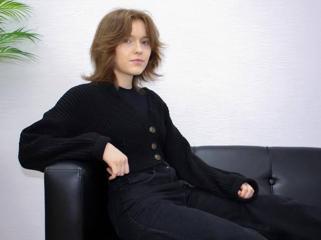 YukiKendal with medium brown wavy hair, black cardigan with buttons, black shirt, and black pants, sitting on black leather couch, looking slightly to the side.