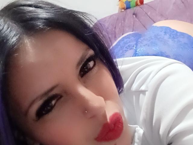NicoleEmiller mit lila Haaren, rotem Lippenstift, schwarzem Eyeliner, Nasenpiercing und weißem Hemd, liegt im Bett in blauen Spitzenunterhosen und zeigt im Hintergrund ihren Hintern..