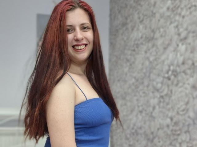 SageJordan a les cheveux roux, un joli sourire, et porte un haut bleu à bretelles fines. Ses seins sont petits, et elle se tient de côté avec une expression joyeuse..