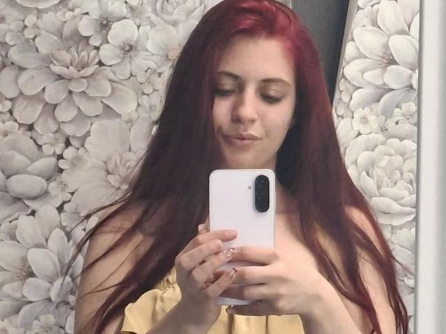SageJordan, con i capelli rossi, sta scattando un selfie con un telefono bianco, indossando una maglietta gialla con una spalla scoperta, e guarda verso il basso con un leggero sorriso. Il seno non è visibile..