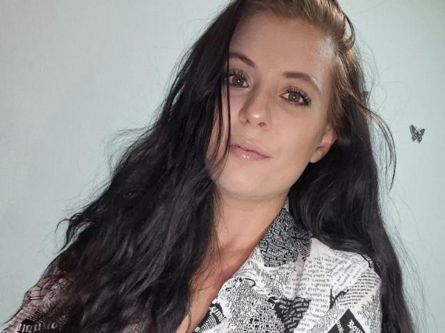 SageJordan tem cabelos longos e pretos, pele clara, olhos castanhos e está vestindo uma camisa estampada com texto de jornal. Ela tem um sorriso discreto e uma pequena tatuagem de borboleta..