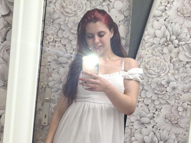 SageJordan, con cabello rojo, vestido blanco con un hombro descubierto, sosteniendo un teléfono, con el flash de la cámara encendido, mostrando senos pequeños, con una sonrisa suave, con papel tapiz floral en el fondo, piel clara, selfie frente al espejo..
