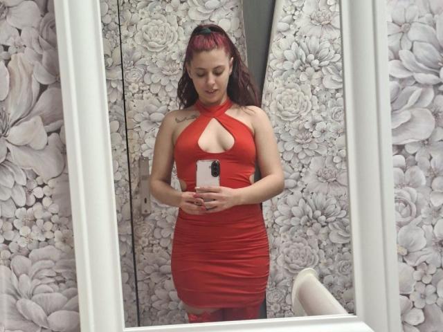 SageJordan met rood haar, gekleed in een rode jurk met een uitsparing, staat voor een spiegel, houdt een telefoon vast en toont haar borsten en een kleine tatoeage op haar rechter schouder..