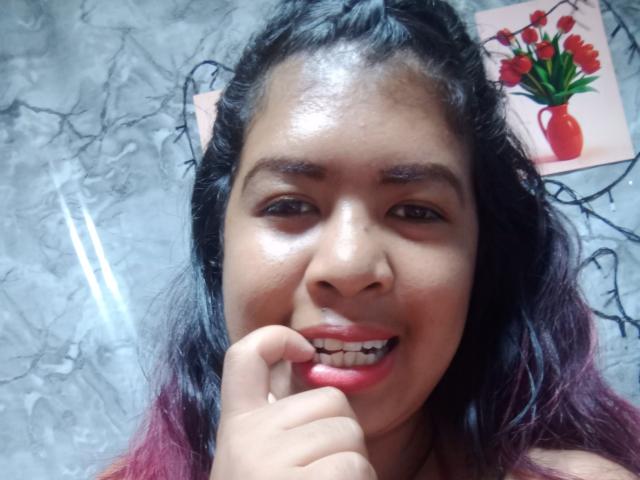 MariahSquirting69 has shiny brown skin, dark curly hair, and is smiling with her finger near her red-lipped mouth. Her nose is small, and her eyebrows are thick.