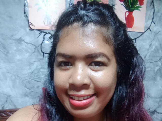 MariahSquirting69 with shiny brown skin, black and purple ombre hair, dark eyebrows, pink lipstick, and a smile, looking at the camera. Her face is centered in the image.