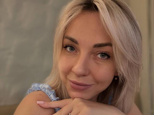 RoseGloss, a blonde woman with light blue eyes, leans her chin on her hand. She has a soft smile, full lips, and subtle makeup. Her light blonde hair is straight, and she wears a blue, frilled, sleeveless top. Her eyebrows are well-groomed, and her long eyelashes enhance her eyes. The image has a warm, slightly blurry effect, highlighting her relaxed, confident expression. RoseGloss's light skin contrasts with her top, and her nails are manicured.