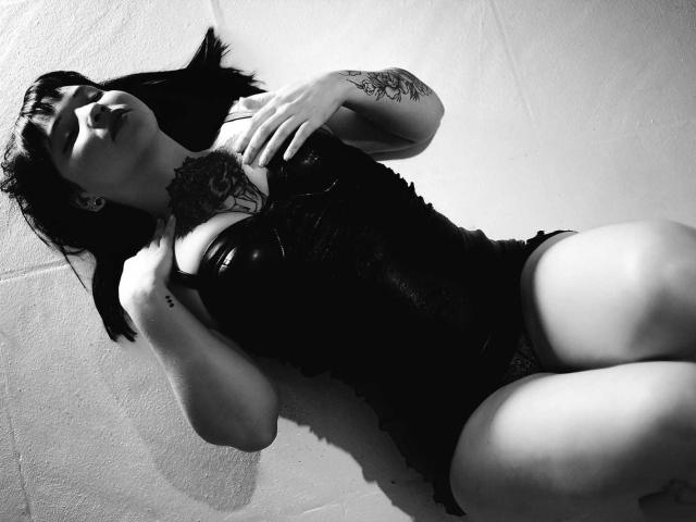 Schwarz-weißes Foto von AlinaMatei, die liegt, ein Spitzenkorsett trägt, Tätowierungen auf ihrem Arm zeigt, mit den Händen auf der Brust, Beine gespreizt, dunkles Haar, geschlossene Augen, leicht geöffneter Mund..