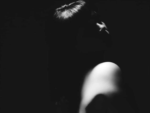 Foto em preto e branco do rosto e ombro parcialmente iluminados de AlinaMatei, com o cabelo preso em um coque, uma sombra escura cobrindo a maior parte de seus traços, destacando seu nariz e ombro..