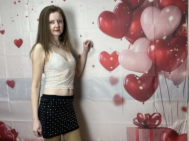 MeganPirs with long brown hair, wearing a white lace crop top and black polka-dot skirt, standing, showing her small tits, against a red balloon and heart wall.