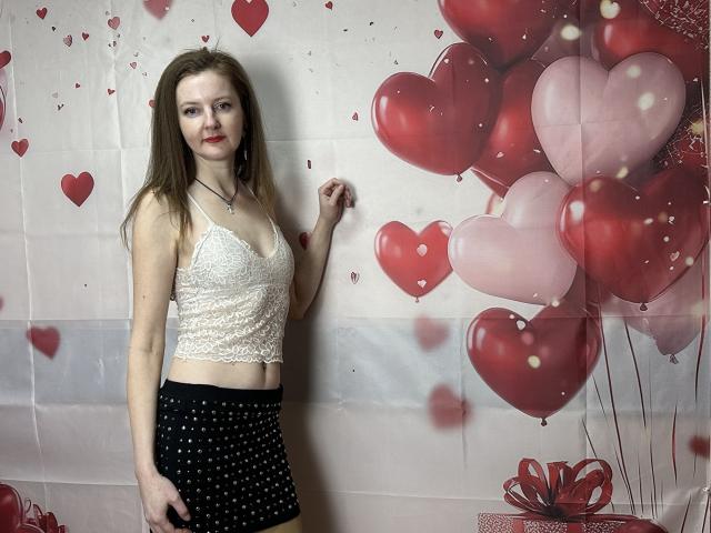 MeganPirs with long brown hair, small tits in a white lace crop top, black polka-dot skirt, standing, smiling, holding her hair, red heart balloons in the background.