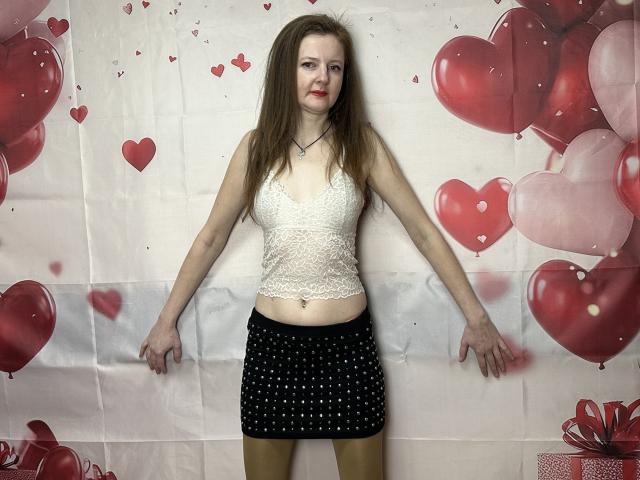 MeganPirs with long brown hair, wearing a white lace crop top and black polka-dot skirt, standing with arms out, showing her tits and small ass against a red balloon backdrop.
